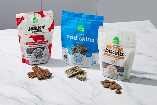 3 bags of open farms dog treats with samples of the treats sitting in front of each bag on a white marble countertop the first bag is red jerky, the second bag is blue cod skins, the third bag is orange better biscuits